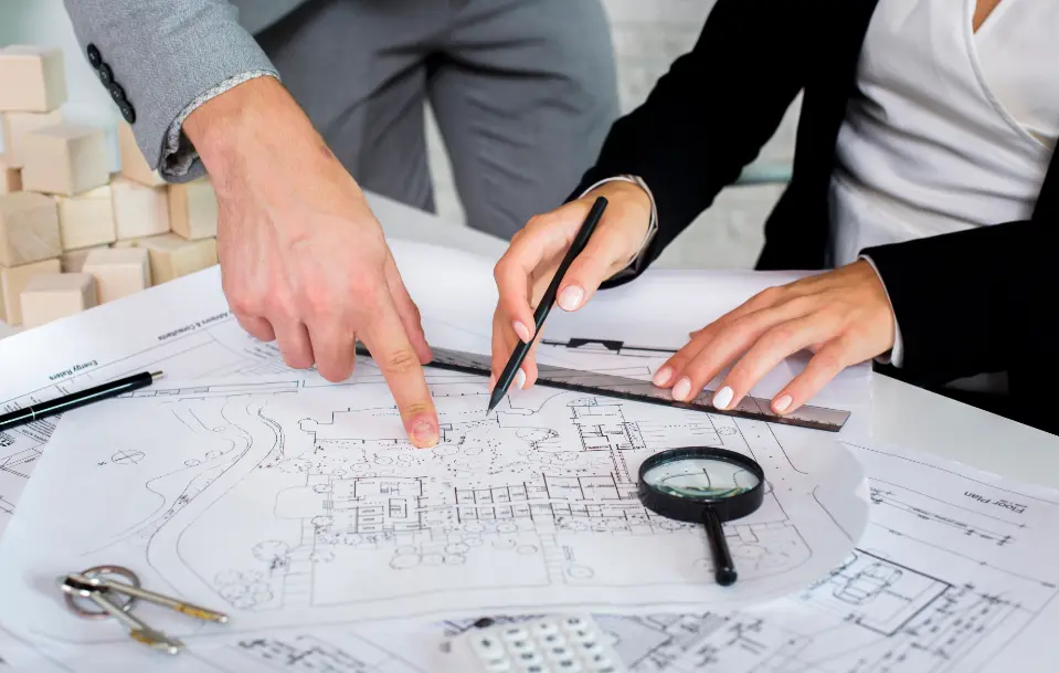 Gros plan sur des mains de professionnels examinant un plan d'architecte avec règle, stylo et loupe, dans le cadre d'un projet de construction ou de rénovation. Baticonnect à Moulins